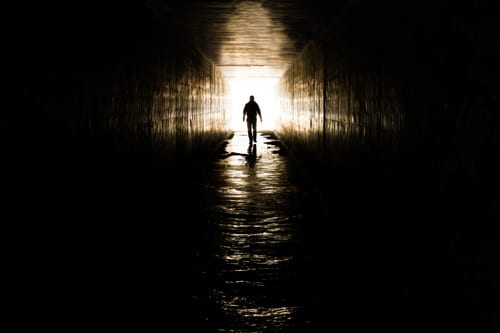 Man walking toward a cross formed by light reflecting off the grungy walls of an underground culvert.